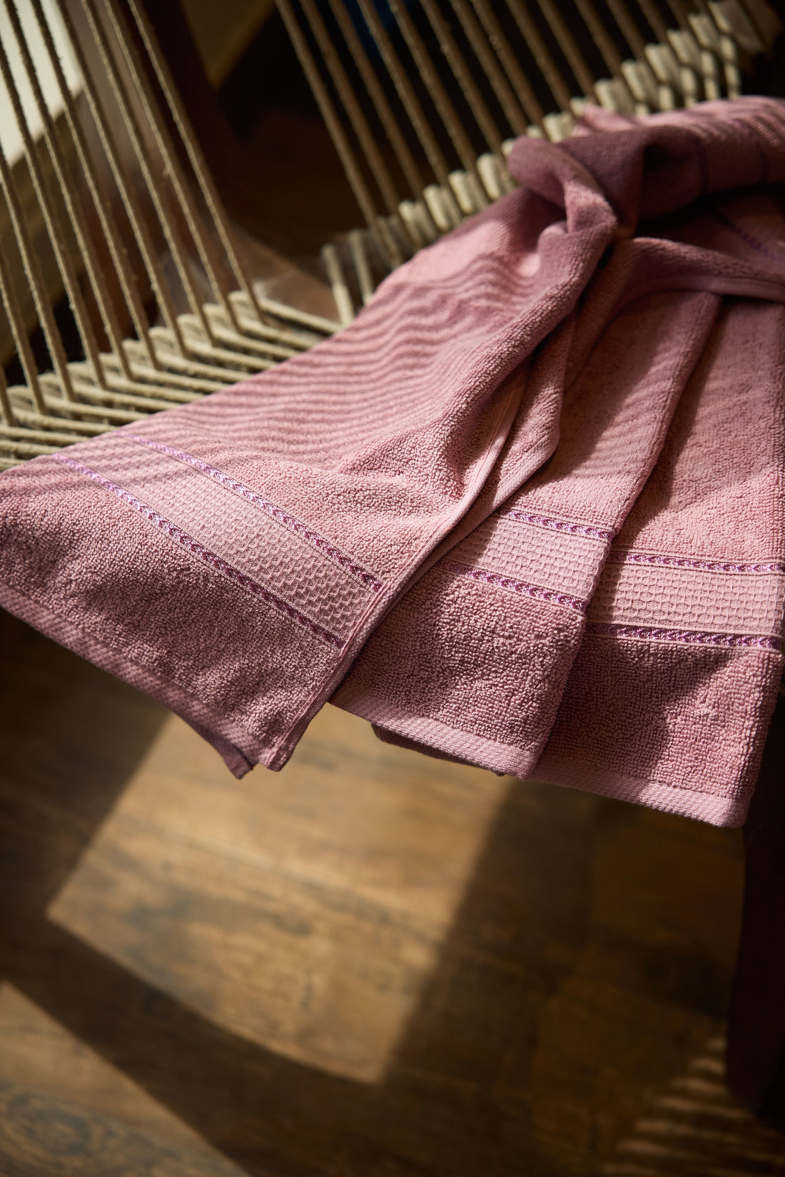 Pink Hyluxe bath towels draped over a wooden chair in warm natural sunlight.