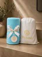 A turquoise blue towel wrapped with a white ribbon and tag stands beside a white drawstring storage bag printed with the Hyluxe logo, set on a beige stone surface in a warm interior setting.