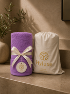 A purple towel roll tied with a cream ribbon and tag, standing beside a cream drawstring storage bag printed with the Hyluxe logo, set on a beige stone surface against a warm beige wall.
