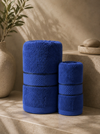 Two rolled blue towels, one large and one small, displayed on a beige stone surface with a textured beige wall, a vase with greenery, and a stone dish in the background.