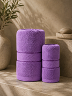 Two purple towels, one larger and one smaller, standing upright on a beige stone surface with a textured wall and decorative vase in the background.