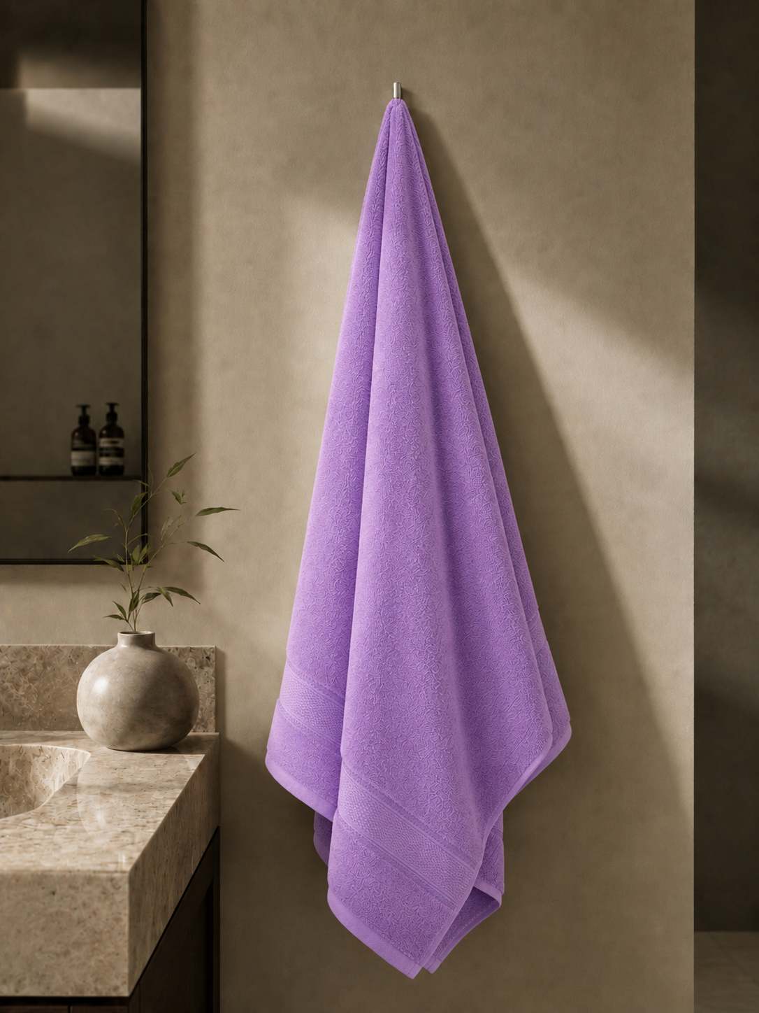 A lavender purple bath towel hangs from a metal hook on a textured beige wall in a minimalist bathroom setting with a stone sink and a small vase.