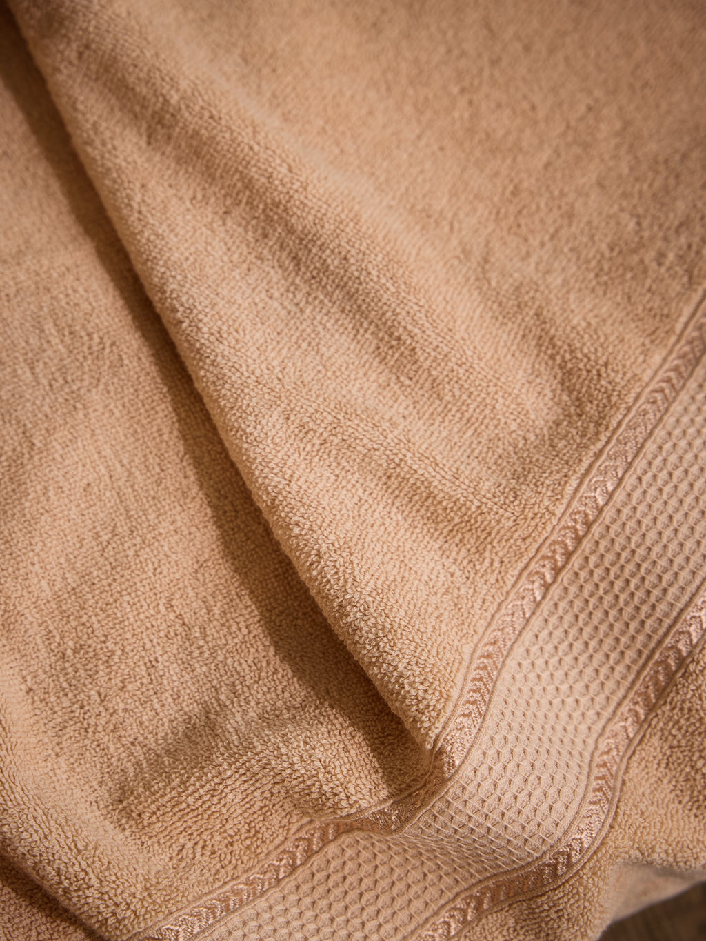 A close-up of a soft beige cotton towel with a textured waffle-weave border and folded drapery.