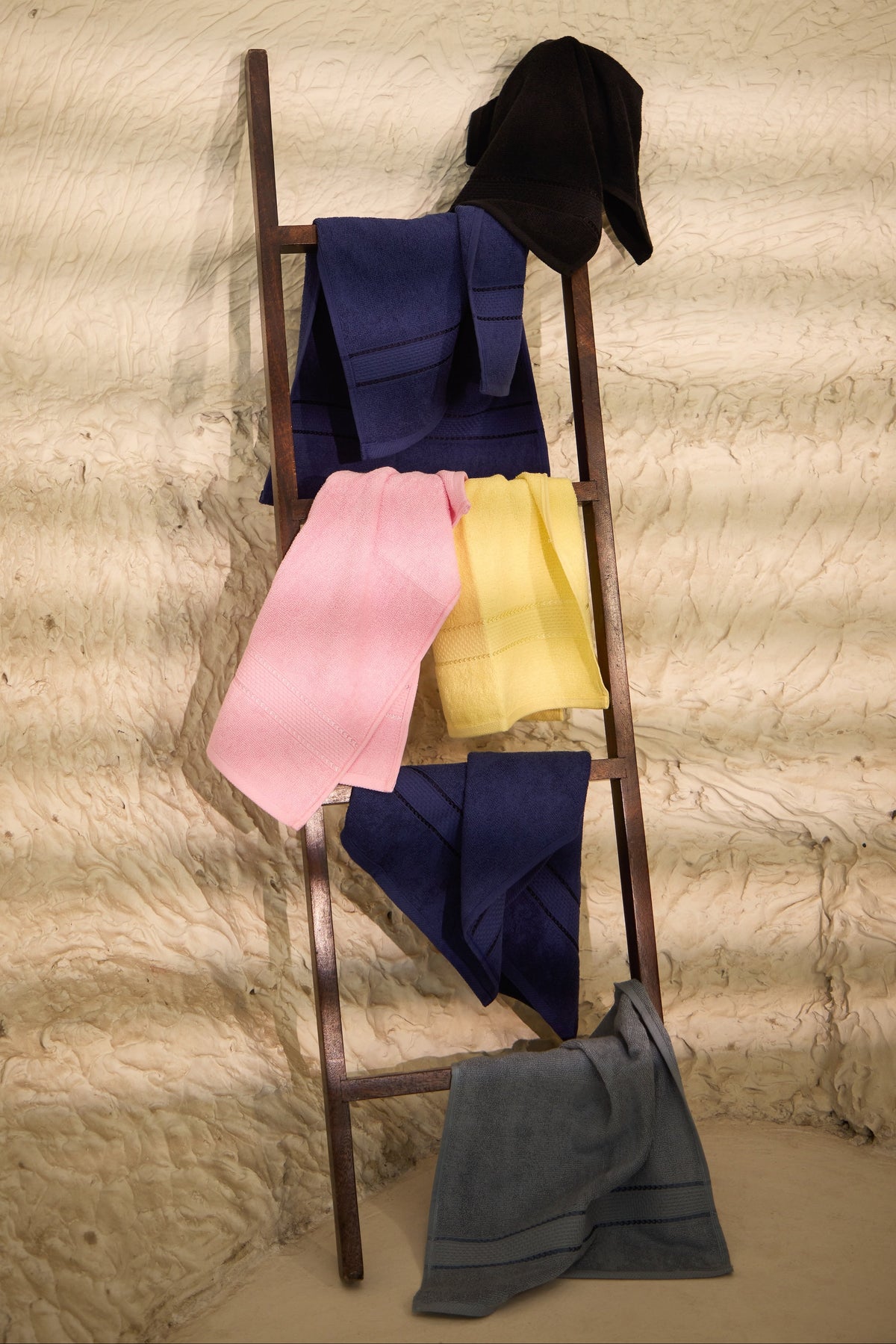 A wooden ladder draped with multiple folded fabric pieces in navy, pink, yellow, black, and gray, set against a textured beige wall.